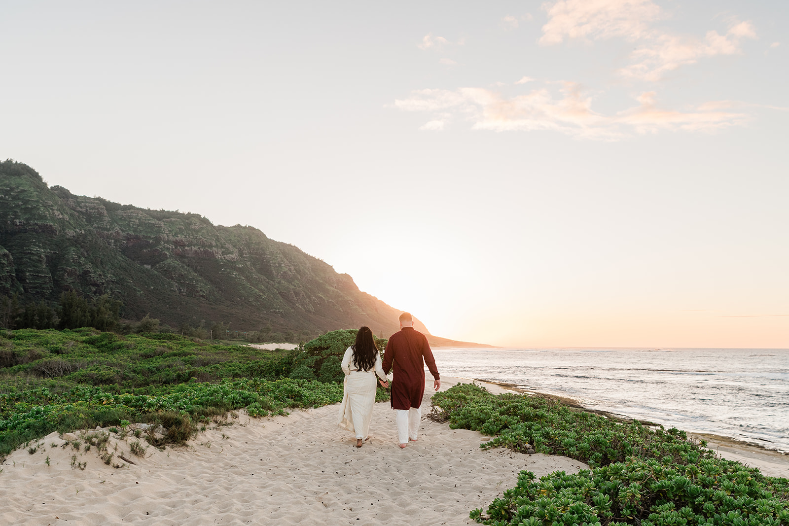 Samira and Michael walking hand-in-hand into the sunset on a beach with mountains in the background during their all-inclusive elopement in Hawaii